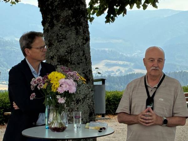 Zwei Männer im Gespräch vor Publikum im Freien an einem Stehtisch mit Blumen, im Hintergrund der Stamm einer Linde und Aussicht auf den Schwarzwald.